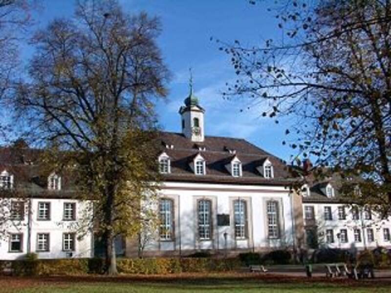 Ein historisches Gebäude mit weißen Wänden und einem kleinen Turm, umgeben von Bäumen mit kahlen Ästen an einem sonnigen Tag.