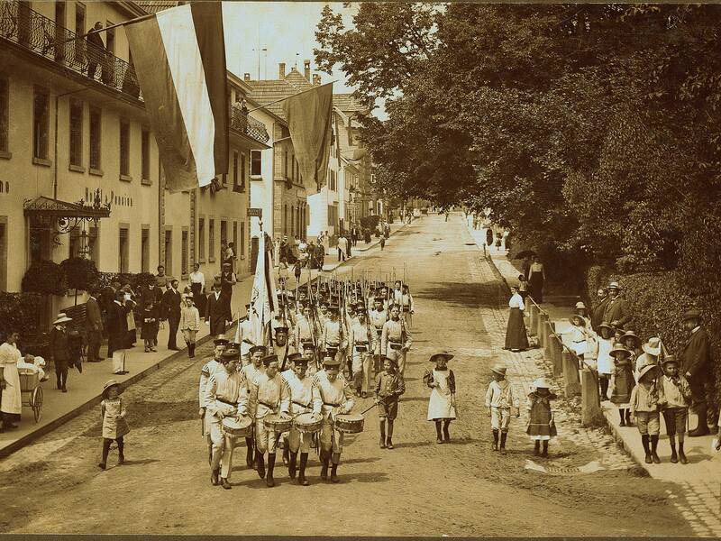 Eine Gruppe von Menschen, darunter Männer in Uniform, marschiert eine Straße entlang, während Zuschauer, einschließlich Kinder, am Rand stehen.
