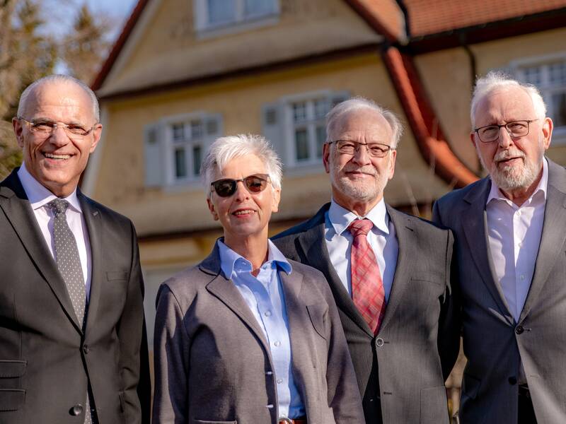 Eine Gruppe von vier lächelnden Personen, drei Männer und eine Frau in Business-Kleidung, steht vor einem Gebäude.