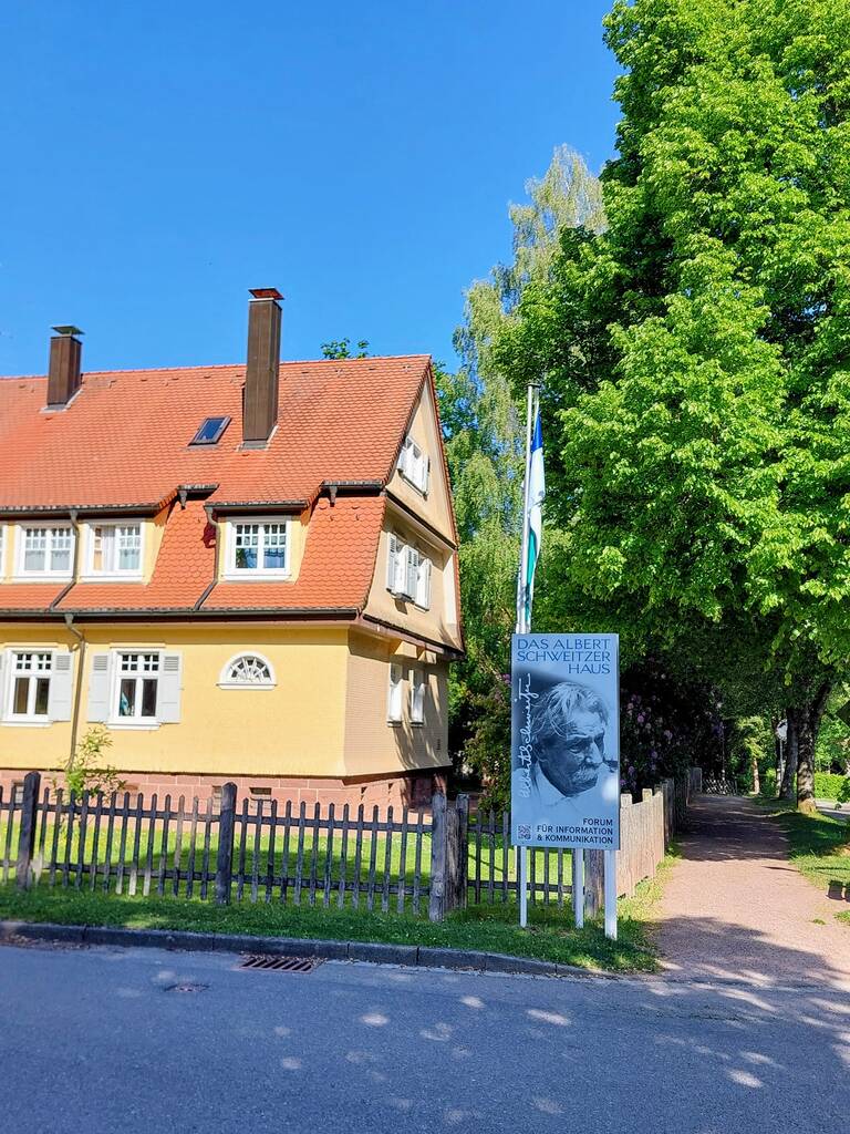 Ein zweigeschossiges gelbes Haus mit rotem Dach steht neben einem Holzzaun und einem Schild mit der Aufschrift "Das Albert Schweitzer Haus", umgeben von einem grünen Baum und blauem Himmel.