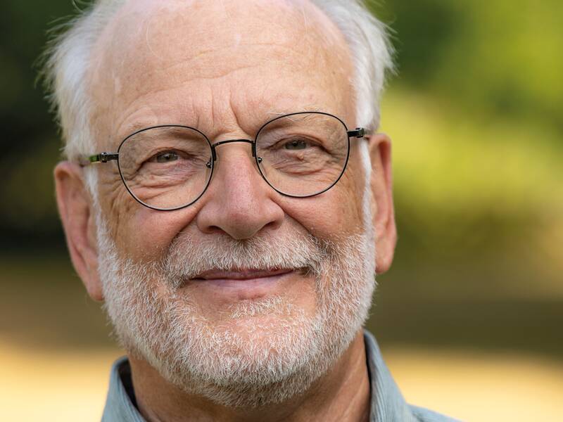 Ein älterer Mann mit Brille und Bart lächelt in die Kamera, während er sich vor einem natürlichen, unscharfen Hintergrund befindet.