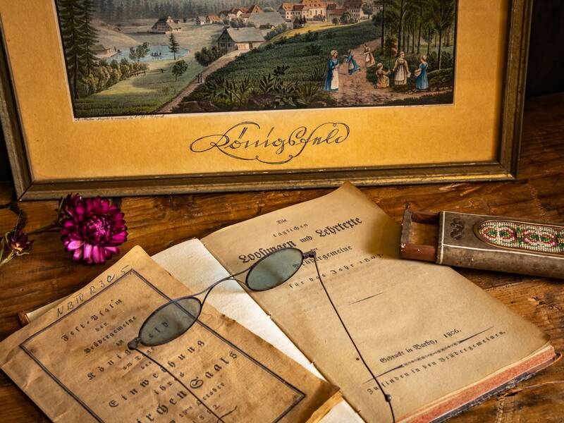Ein altes, eingerahmtes Bild mit einer idyllischen Landschaft steht auf einem Holztisch, umgeben von historischen Dokumenten, einer Brille, einem dekorativen Metallbehälter und einer roten Blüte.