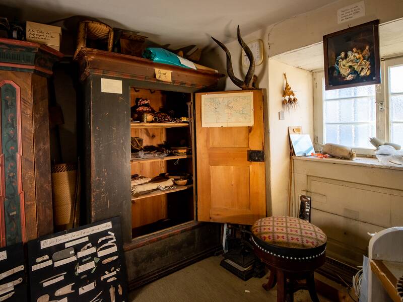 Ein rustikaler Raum mit einem geöffneten Schrank voller alter Gegenstände, einem Hocker, einem Fenster mit etwas Dekoration und verschiedenen Schautafeln, die Messer präsentieren.