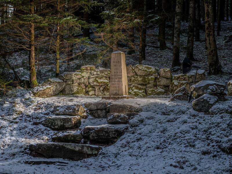 Ein schneebedeckter Wald mit einer Gedenktafel auf einem kleinen Steinsockel, umgeben von einer niedrigen Steinmauer.