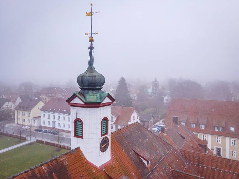 Der obere Teil eines Kirchturms mit einer Wetterfahne ist vor einem nebligen Hintergrund mit mehreren Häuserdächern zu sehen.