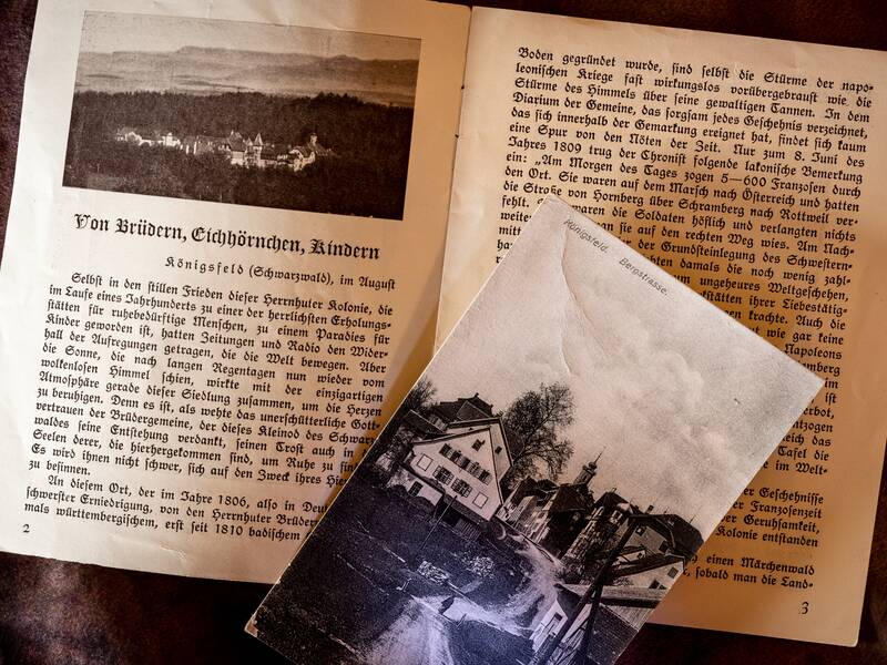 Ein aufgeschlagenes Buch mit einem Schwarz-Weiß-Foto von Gebäuden und Text über „Von Brüdern, Eichhörnchen, Kindern“ sowie eine Postkarte liegt darauf.