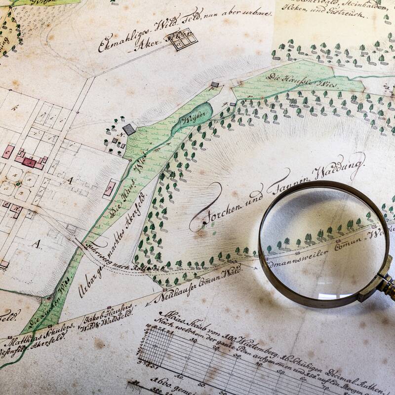 Eine historische Landkarte zeigt Felder, Gebäudepläne und topografische Details, über die eine Lupe liegt.