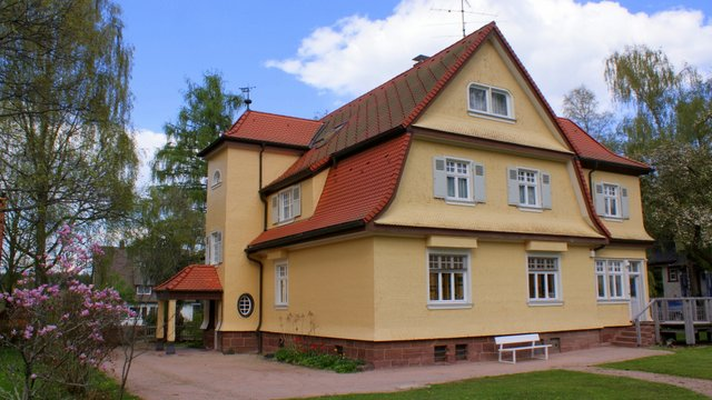 Ein zweistöckiges, gelbes Haus mit rotem Dach und weißen Fensterrahmen steht inmitten eines grünen Gartens mit Bäumen und blühenden Sträuchern. Albert-Schweitzer-Haus