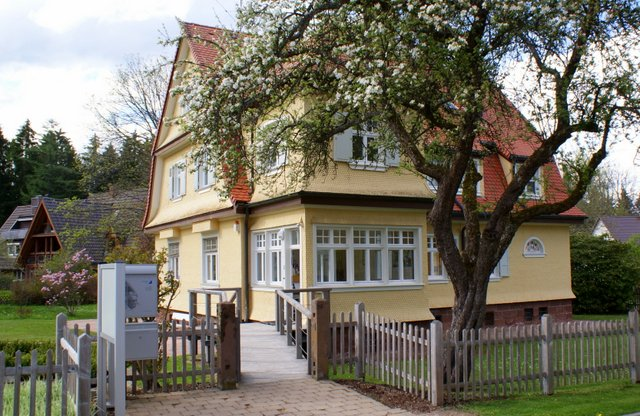 Ein zweistöckiges, gelbes Haus mit rotem Dach und zahlreichen Fenstern steht hinter einem Holzzaun, neben einem blühenden Baum.
