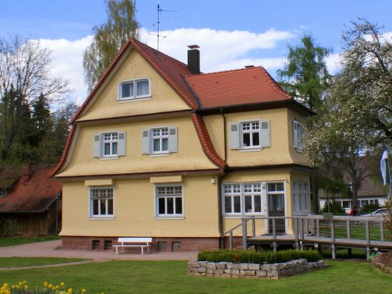 Ein zweistöckiges gelbes Haus mit rotem Dach steht in einem gepflegten Garten mit blühendem Baum und Rasen. Foto zeigt das Albert Schweitzer Haus