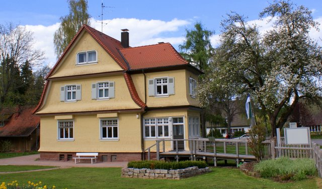 Ein zweistöckiges gelbes Haus mit rotem Dach steht in einem gepflegten Garten mit blühendem Baum und Rasen. Foto zeigt das Albert Schweitzer Haus
