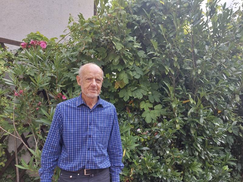 Ein älterer Mann in einem blau karierten Hemd steht vor einem üppigen grünen Busch mit rosa Blüten.