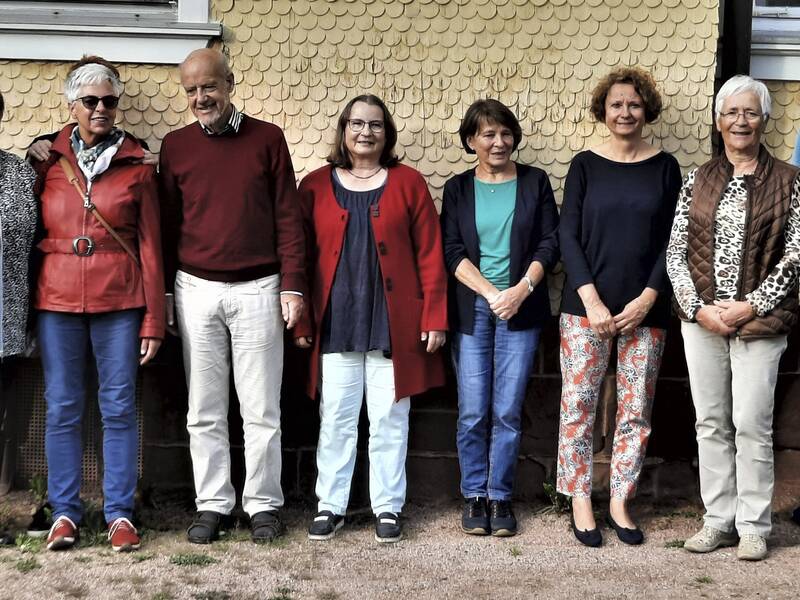 Eine Gruppe von zehn Personen steht lächelnd nebeneinander vor einer gelben Hauswand.