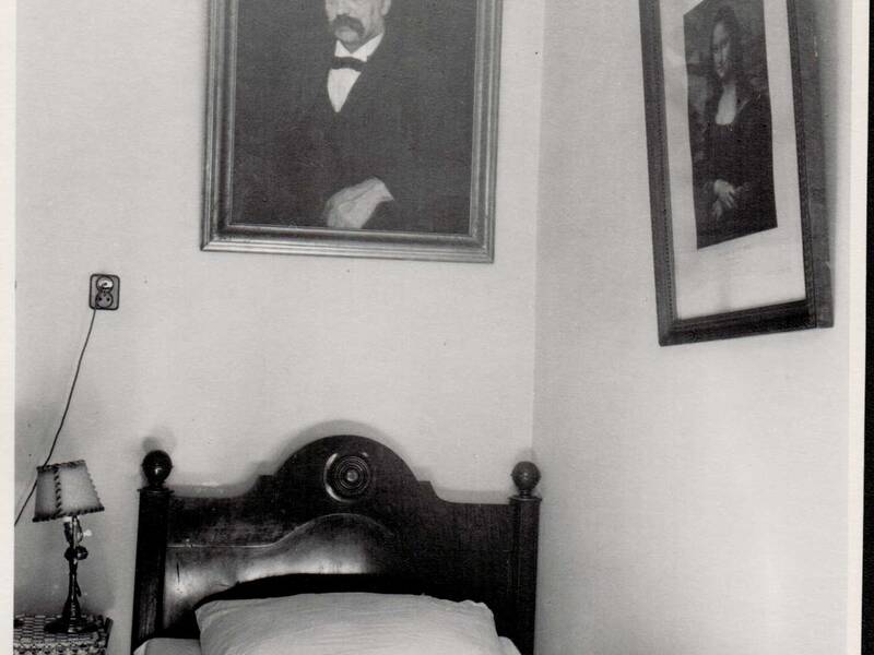 Ein schmales Bett mit einer geschnitzten Holzlehne steht unter zwei an der Wand hängenden Porträtbildern.