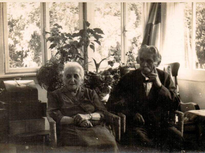 Ein älteres Ehepaar sitzt in einem Wohnzimmer vor großen Fenstern und Pflanzen.