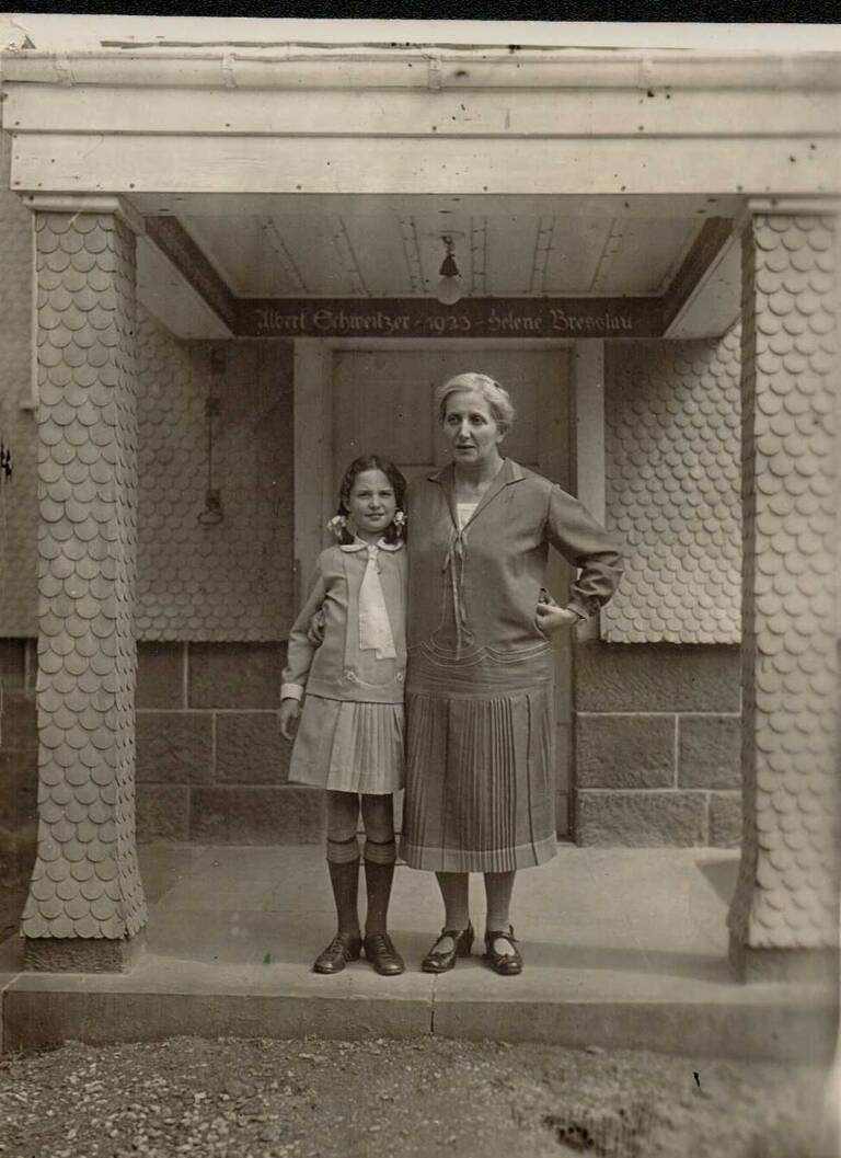 Eine Frau und ein Mädchen stehen auf einer Veranda vor einer Tür, wobei die Frau den Arm um das Mädchen legt.