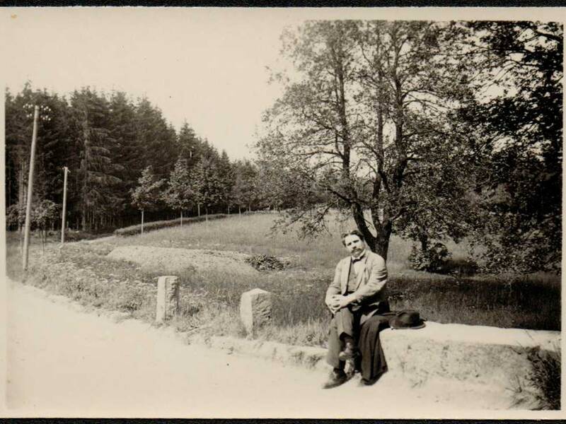 Ein Mann in Anzug und Hut sitzt auf einer Steinmauer neben einer Landstraße mit Bäumen und Wiese im Hintergrund.
