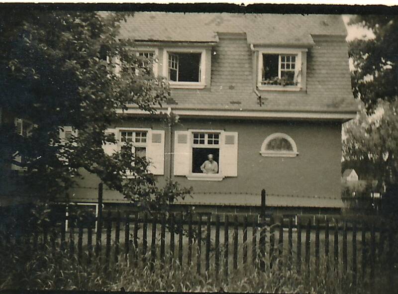 Ein zweistöckiges Haus mit geöffnetem Fenster im Erdgeschoss, in dem eine Person sichtbar ist, umgeben von Bäumen und einem Zaun im Vordergrund.