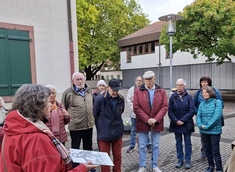 Eine Gruppe von älteren Menschen steht draußen in einem Kreis, während eine Person ein Bild in der Hand hält und zu ihnen spricht.