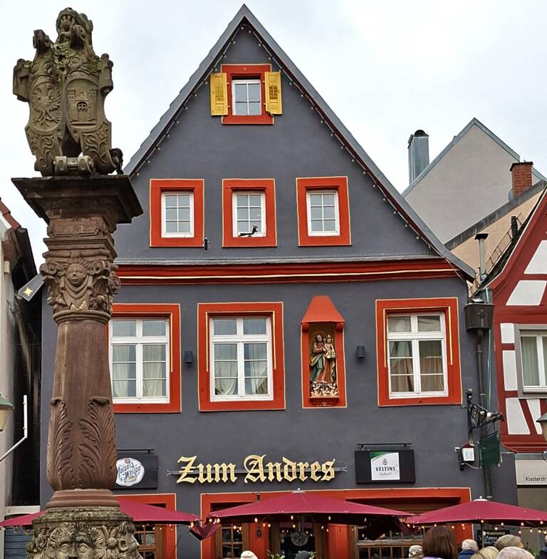 Eine belebte Straßenszene zeigt Menschen vor einem traditionellen Gebäude mit dunkler Fassade und orangefarbenen Fensterrahmen sowie einem aufwendig verzierten Brunnen im Vordergrund.