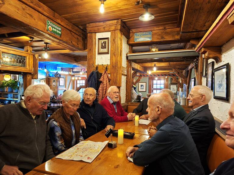 Eine Gruppe älterer Menschen sitzt um einen Holztisch in einem rustikalen Restaurant und unterhält sich, während eine brennende Kerze in der Mitte des Tisches steht.