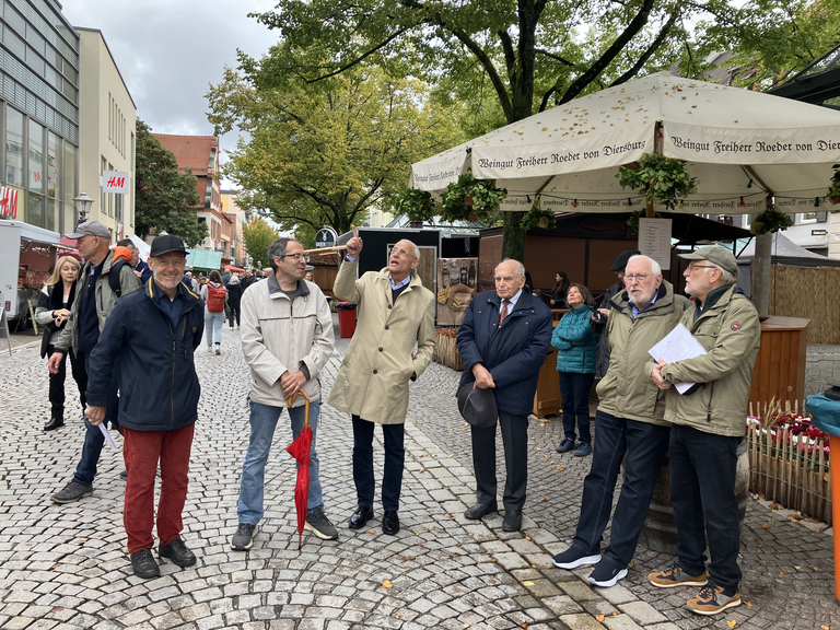 Eine Gruppe von älteren Menschen steht unter einem Pavillon auf einem gepflasterten Platz, während ein Mann auf etwas zeigt.