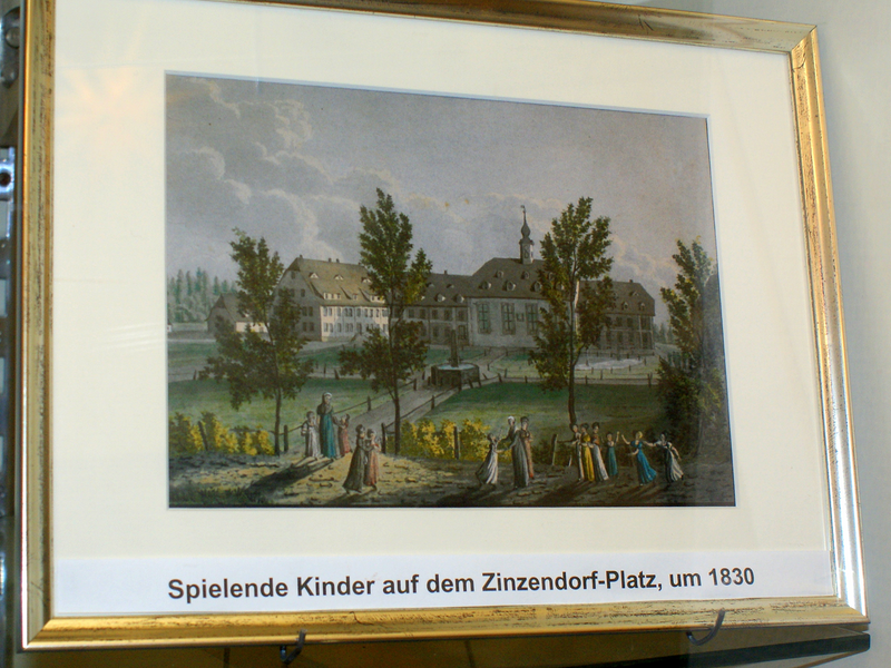 Ein historisches Gemälde zeigt spielende Kinder auf einem Platz vor einem großen Gebäude, umringt von Bäumen, beschriftet mit "Spielende Kinder auf dem Zinzendorf-Platz, um 1830".