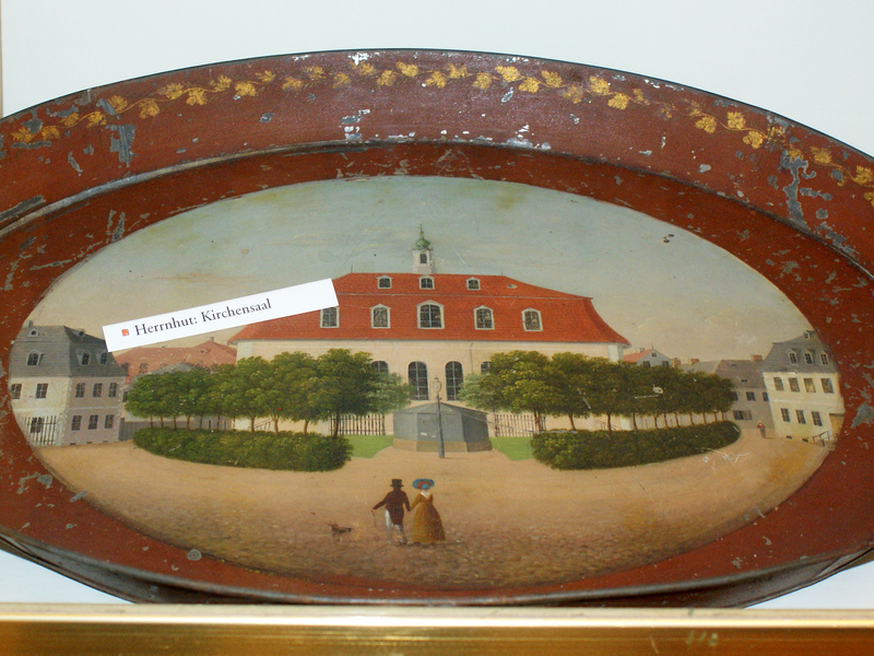 Ein ovales Tablett zeigt ein gemaltes Bild eines Gebäudes mit roten Dach und Bäumen im Vordergrund, beschriftet als "Herrnhut: Kirchensaal".