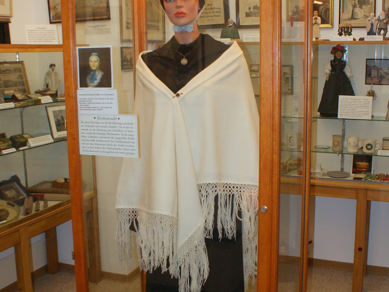 Eine Schaufensterpuppe in einem gläsernen Schaukasten trägt traditionelle Kleidung und einen weißen Schal, umgeben von historischen Fotos und Objekten.