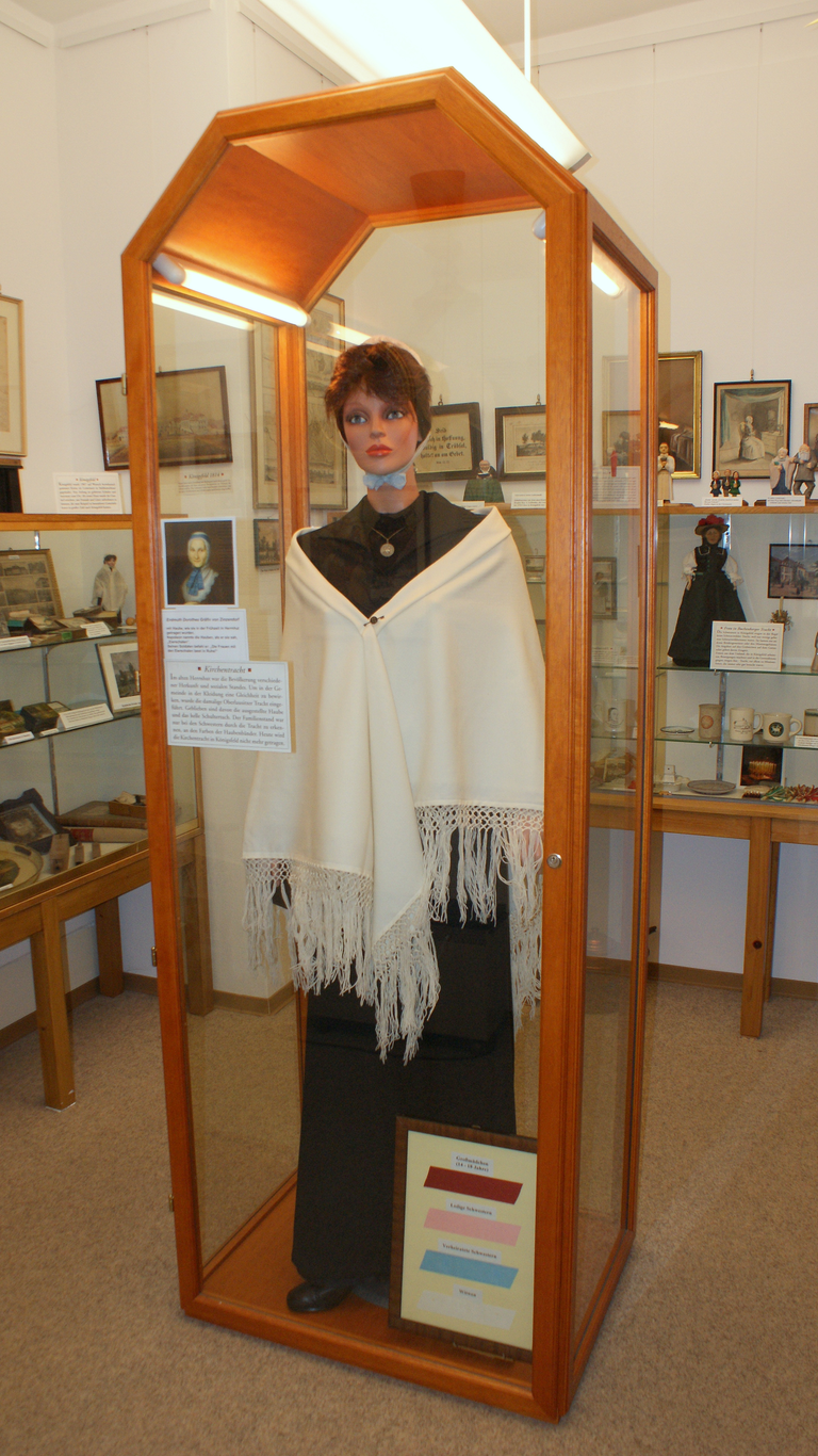 Eine Schaufensterpuppe in einem gläsernen Schaukasten trägt traditionelle Kleidung und einen weißen Schal, umgeben von historischen Fotos und Objekten.
