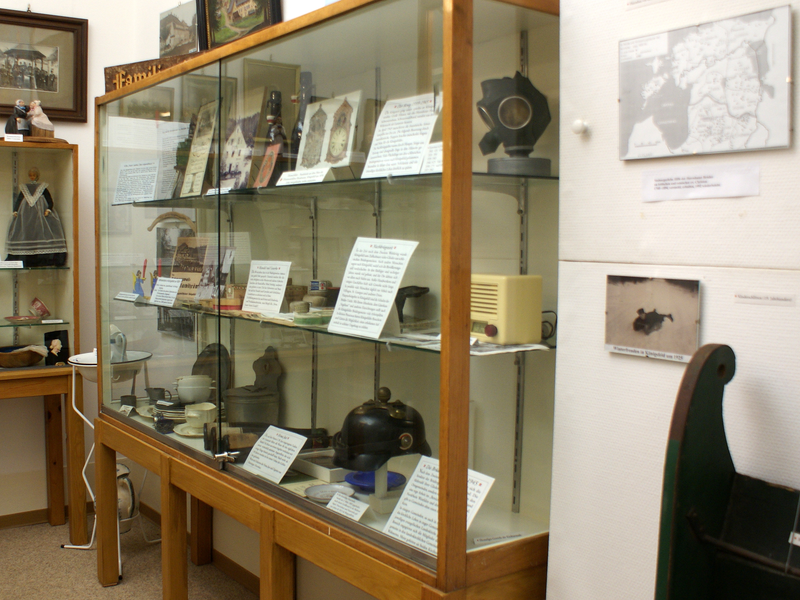 In einem Museum zeigt eine Glasvitrine eine Sammlung historischer Objekte und Dokumente, während an der Wand eine Landkarte und ein Foto hängen.