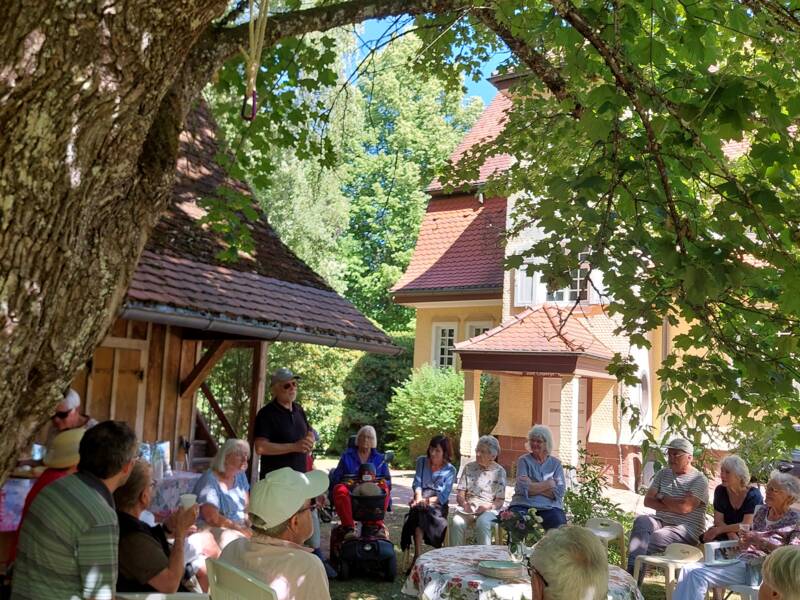 Eine Gruppe von Menschen sitzt unter einem großen Baum im Schatten und hört einer Person zu, die vor einem Haus spricht.