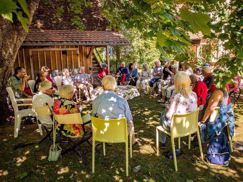 Bei einer Veranstaltung im Sommer sitzen im Garten Menschen unter hohen Bäumen