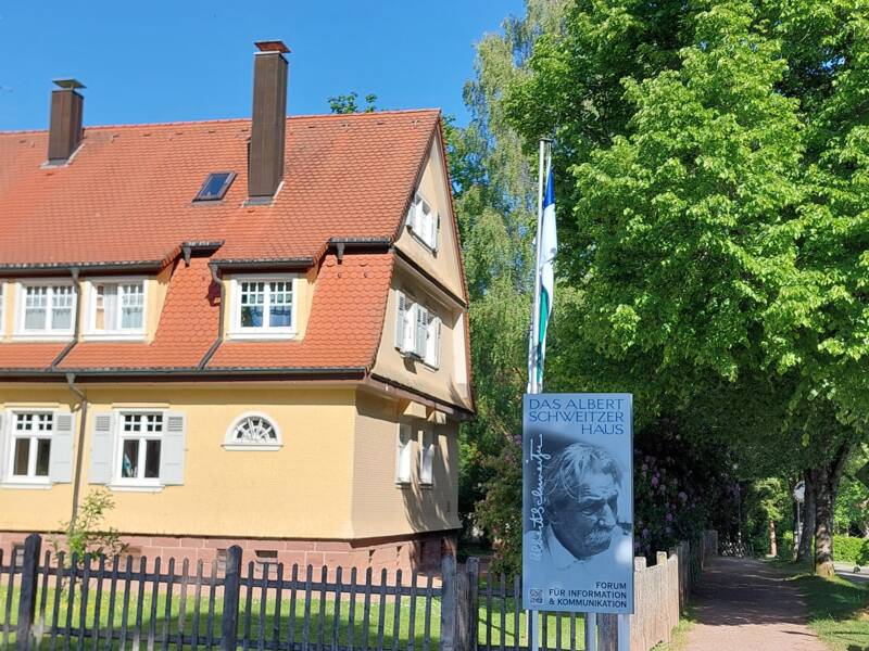 Ein zweistöckiges gelbes Haus mit rotem Dach steht neben einem Schild, das auf das Albert-Schweitzer-Haus hinweist, an einem sonnigen Tag vor einem blauen Himmel und grünen Bäumen.