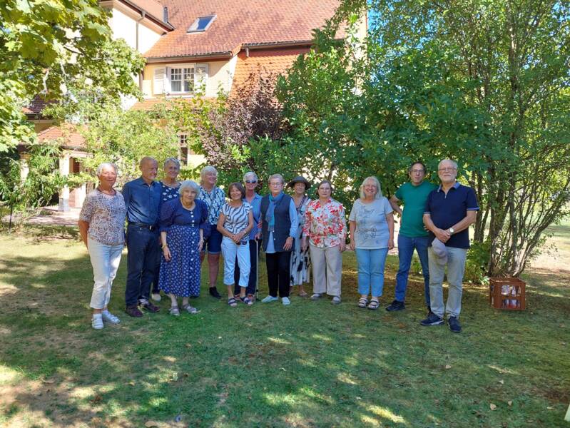 Eine Gruppe sommerlich gekleideter Menschen steht versammelt unter Bäumen im Garten des Albert Schweitzer Hauses. Warmes Licht fällt durch die Blätter und Sträucher auf die Menschen und den Rasen