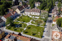 Das Bild zeigt eine Luftaufnahme eines symmetrisch angelegten Parks mit Rasenflächen, Wegen und einem großen Gebäude im Hintergrund, umgeben von Bäumen und angrenzenden Straßen.